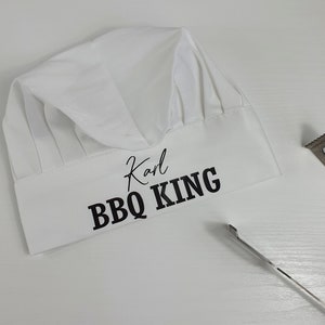 Pu&ograve; includere: Un cappello da chef bianco con la scritta "Karl BBQ KING" in nero. Il cappello presenta un classico design a pieghe ed &egrave; realizzato in un tessuto morbido. Una spatola di metallo &egrave; visibile sullo sfondo. Ideale per gli amanti del barbecue.