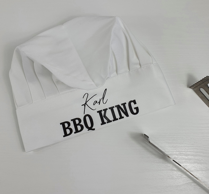 Pu&ograve; includere: Un cappello da chef bianco con la scritta "Karl BBQ KING" in nero. Il cappello presenta un classico design a pieghe ed &egrave; realizzato in un tessuto morbido. Una spatola di metallo &egrave; visibile sullo sfondo. Ideale per gli amanti del barbecue.