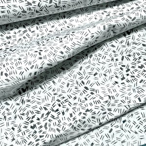 May include: White fabric with a repeating pattern of black dashes and short lines. The fabric is folded, creating shadows and texture. The design is simple and modern, suitable for various sewing and crafting projects.
