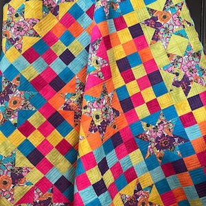 May include: A colorful quilt with a patchwork design. The quilt features a vibrant mix of colors, including pink, orange, yellow, blue, and purple. Large star shapes with floral patterns are interspersed throughout the quilt's square pattern.
