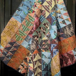 May include: A colorful patchwork quilt with a variety of geometric patterns. The quilt features squares and triangles in shades of blue, orange, green, and brown. The quilt is draped over a dark surface, showcasing its intricate design.