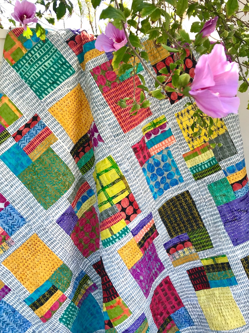 May include: A colorful patchwork quilt with a geometric pattern. The quilt is made of various fabrics in shades of blue, green, yellow, red, and purple. The quilt is draped over a white surface with a branch of pink flowers in the background.