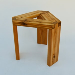 May include: A triangular wooden stool with three legs. The stool is made of natural wood with a light brown finish.
