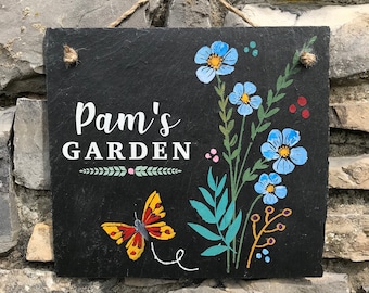 May include: A black slate garden sign with the text "Pam's GARDEN" in white script. The sign features a painted butterfly and blue flowers with green leaves. A twine loop is attached for hanging.