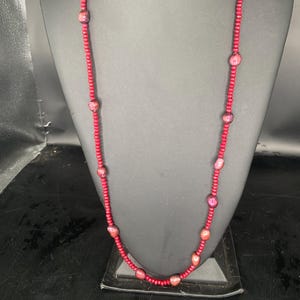 May include: A long, red beaded necklace with larger, oval-shaped beads. The necklace is displayed on a black mannequin, highlighting its length and design. The beads have a slightly iridescent quality, adding visual interest.