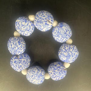 以下が含まれることがあります： 青と白の模様が入った、大きくて丸いビーズのブレスレット。このブレスレットは、模様入りのビーズと、小さく無地の白いビーズを交互に配置しています。ビーズは繋がれて円形を形成しています。