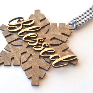 May include: A wooden snowflake ornament with the word "Blessed" in cursive script. The snowflake is stained a dark brown color, and the word "Blessed" is a lighter wood tone. A black and white checkered ribbon is attached for hanging.