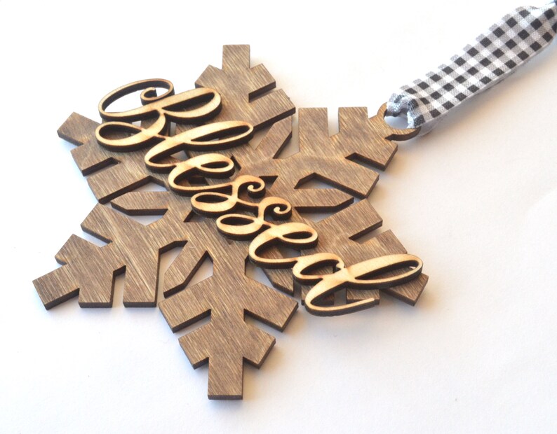 May include: A wooden snowflake ornament with the word "Blessed" in cursive script. The snowflake is stained a dark brown color, and the word "Blessed" is a lighter wood tone. A black and white checkered ribbon is attached for hanging.