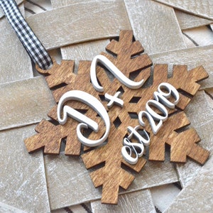 May include: Wooden snowflake ornament with the letters "D" and "N" and the text "est 2019" in white. The snowflake is brown with a black and white checkered ribbon for hanging.