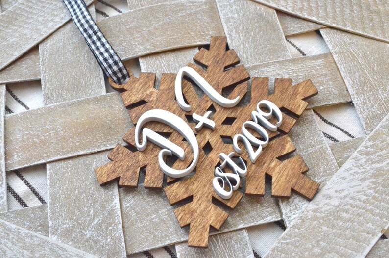 May include: Wooden snowflake ornament with the letters "D" and "N" and the text "est 2019" in white. The snowflake is brown with a black and white checkered ribbon for hanging.