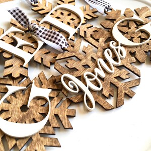 May include: Wooden snowflake ornaments with white letters and the name "Jones" in cursive. The snowflakes are brown with a wood grain pattern, and the letters are white. Each ornament has a black and white checkered ribbon for hanging.