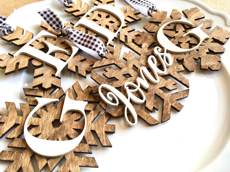 May include: Wooden snowflake ornaments with white letters and the name "Jones" in cursive. The snowflakes are brown with a wood grain pattern, and the letters are white. Each ornament has a black and white checkered ribbon for hanging.