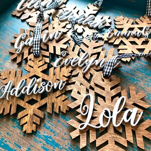 May include: Wooden snowflake ornaments with white cursive names, including "Chloe", "Gavin", "Addison", and "Lola". Each ornament has a black and white checkered ribbon for hanging. The ornaments are displayed on a distressed blue wooden surface.