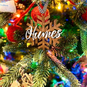 May include: A wooden snowflake Christmas ornament with the name "Humes" in white script. The ornament hangs on a decorated Christmas tree with red ornaments and colorful lights. The snowflake is brown.