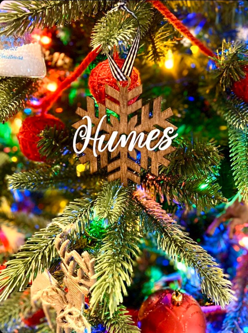 May include: A wooden snowflake Christmas ornament with the name "Humes" in white script. The ornament hangs on a decorated Christmas tree with red ornaments and colorful lights. The snowflake is brown.