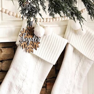 May include: Two white cable-knit Christmas stockings with white pom-poms and a wooden snowflake ornament that reads "Snyder". The stockings are hanging on a fireplace mantel, decorated with greenery and a wooden bead garland.