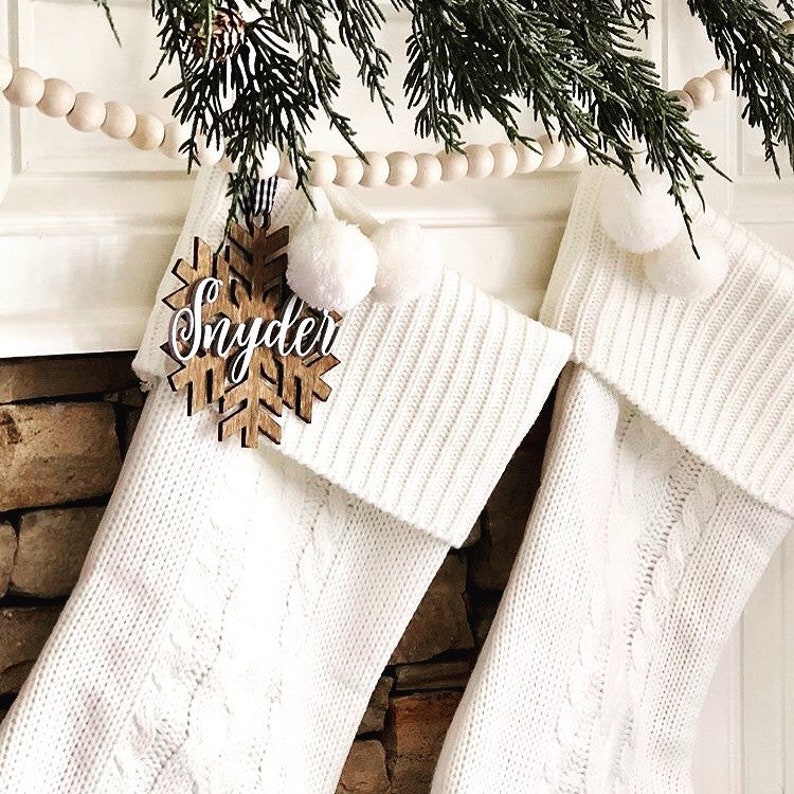 May include: Two white cable-knit Christmas stockings with white pom-poms and a wooden snowflake ornament that reads "Snyder". The stockings are hanging on a fireplace mantel, decorated with greenery and a wooden bead garland.
