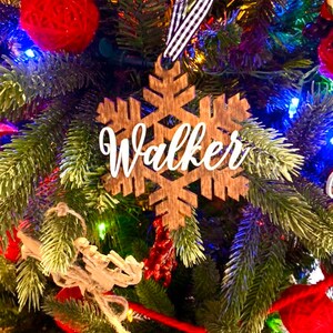 May include: A wooden snowflake Christmas ornament with the name "Walker" in white script. The ornament hangs from a black and white striped ribbon against a backdrop of a decorated Christmas tree with red ornaments and lights.