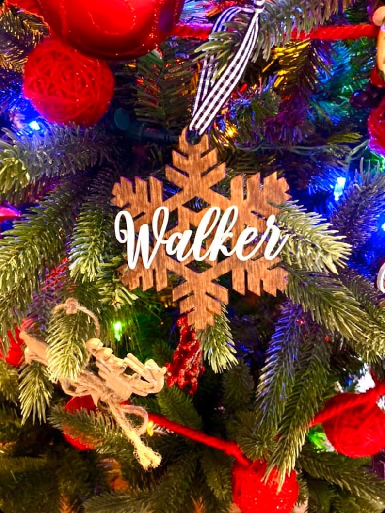 May include: A wooden snowflake Christmas ornament with the name "Walker" in white script. The ornament hangs from a black and white striped ribbon against a backdrop of a decorated Christmas tree with red ornaments and lights.
