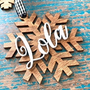 May include: Wooden snowflake ornament with the name "Jola" in white script. The snowflake is made of light brown wood and has a black and white checkered ribbon for hanging. The ornament is on a distressed blue wooden surface.