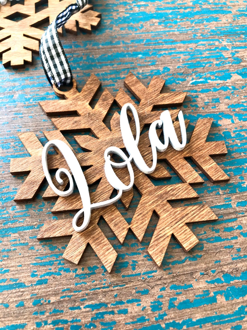 May include: Wooden snowflake ornament with the name "Jola" in white script. The snowflake is made of light brown wood and has a black and white checkered ribbon for hanging. The ornament is on a distressed blue wooden surface.
