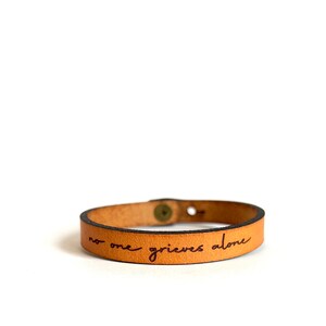 May include: A tan leather bracelet with the words "no one grieves alone" engraved in brown ink.