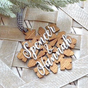 May include: Wooden snowflake ornament with the names "Spencer & Hannah" in white script. The snowflake is made of light brown wood and has a black and white checkered ribbon for hanging. The ornament is on a woven background.