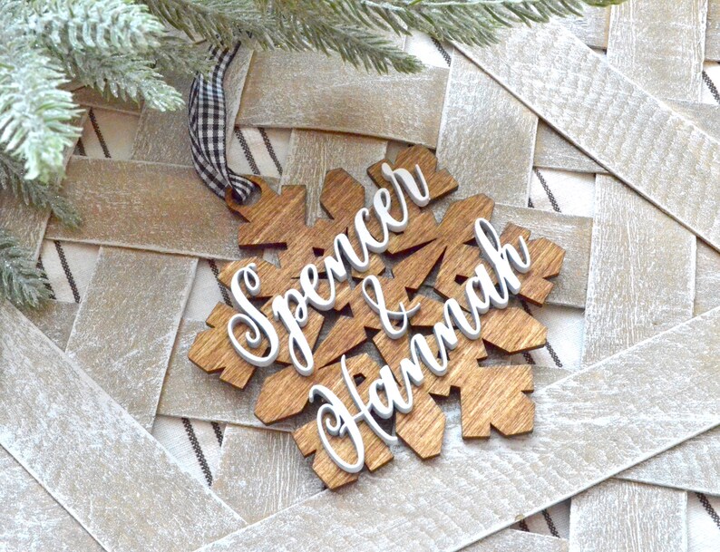 May include: Wooden snowflake ornament with the names "Spencer & Hannah" in white script. The snowflake is made of light brown wood and has a black and white checkered ribbon for hanging. The ornament is on a woven background.