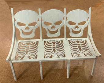 May include: White metal bench with three skull-shaped backrests and ribcage-shaped seats.