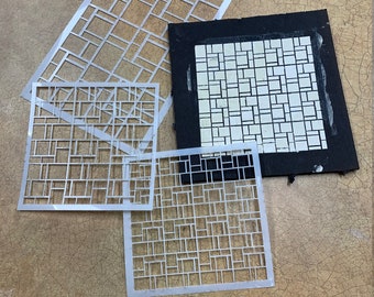 Könnte beinhalten: Vier Schablonen mit einem geometrischen Muster aus Quadraten und Rechtecken. Die Schablonen sind aus weißem Kunststoff und liegen auf einer braunen Oberfläche.