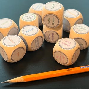 K&ouml;nnte beinhalten: Eine Ansammlung von Holzw&uuml;rfeln mit Aufdrucken wie "Mystery", "Outer Space" und "Time Travel". Ein orangefarbener Bleistift mit der Aufschrift "USA" liegt ebenfalls auf der Oberfl&auml;che.