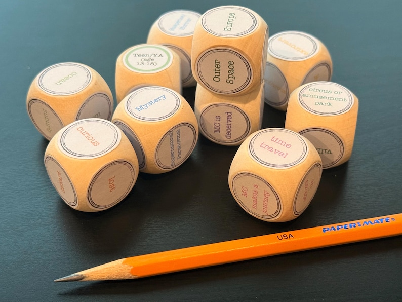 K&ouml;nnte beinhalten: Eine Ansammlung von Holzw&uuml;rfeln mit Aufdrucken wie "Mystery", "Outer Space" und "Time Travel". Ein orangefarbener Bleistift mit der Aufschrift "USA" liegt ebenfalls auf der Oberfl&auml;che.