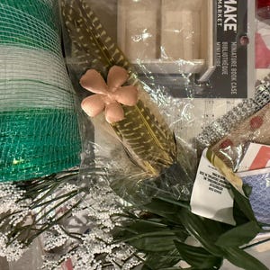 May include: A collection of craft supplies including green mesh ribbon, a feather with a pink flower, artificial white flowers, and a miniature book case. Other items include playing cards and decorative ribbon.