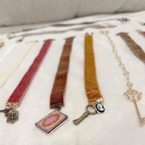 May include: A collection of velvet ribbon bookmarks in various colors, including white, red, and brown. Each bookmark features a decorative charm, such as a key, a cameo, or a miniature book with the text "Little Wishes".
