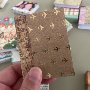 May include: A small, handmade journal with a brown cover featuring gold airplane designs. The spine has a vintage map pattern. Other small journals with patterned covers are in the background.