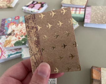 May include: A small, handmade journal with a brown cover featuring gold airplane designs. The spine has a vintage map pattern. Other small journals with patterned covers are in the background.