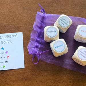 May include: A set of wooden dice with words like "extrovert" and "historical" printed on them, along with a card that says "Children's Book". The dice are in a purple drawstring bag.