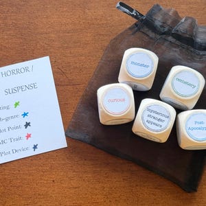 May include: Wooden dice with words like "monster" and "cemetery" are displayed next to a small black bag. A card reads "HORROR / SUSPENSE" with categories like "Setting" and "Plot Point."