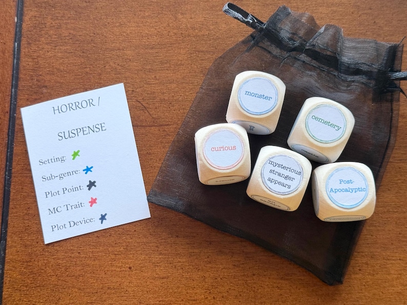 May include: Wooden dice with words like "monster" and "cemetery" are displayed next to a small black bag. A card reads "HORROR / SUSPENSE" with categories like "Setting" and "Plot Point."