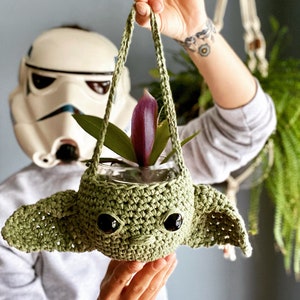 May include: A green crocheted Yoda-shaped hanging planter with a small plant inside. The planter has black eyes and is hanging from a white rope.