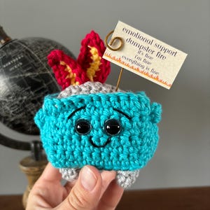 May include: A crocheted blue and gray dumpster fire with a smiling face and red and yellow flames. The fire has a tag that reads "emotional support dumpster fire it's fine i'm fine everything is fine".