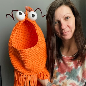 May include: A crocheted orange hanging basket with fringe, featuring large white googly eyes and red pipe cleaner antennae. The basket is being held by a person wearing a tie-dye shirt.