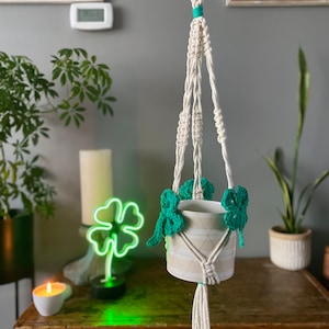 May include: A white macrame plant hanger with a white pot and green crocheted shamrock accents. The hanger is hanging from the ceiling.