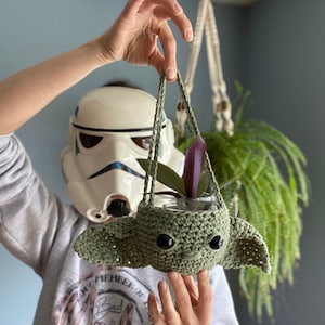 Può includere: Un vaso da fiori in crochet verde a forma di personaggio della saga di Star Wars, che contiene una piccola pianta con foglie viola. Il vaso è appeso a un cordoncino in macramé.