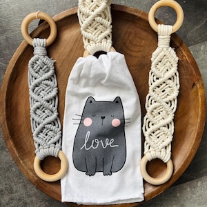 May include: Three white cotton dish towels with a gray cat and the word "love" printed on them. Each towel has a wooden ring and a macrame loop in gray, white, or natural colors.