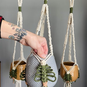 May include: Three macrame plant hangers with green and beige accents. The hangers are made of natural cotton rope and feature a knotted design. The hangers are hanging from a black metal rod. A hand is holding a gray ceramic pot with a geometric pattern.