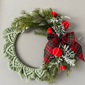 May include: A green macrame wreath with red pom poms and a red and black plaid bow. The wreath is decorated with greenery and sprigs of white dusty miller.