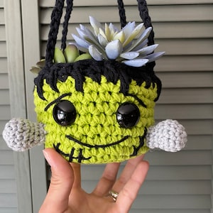May include: A green crocheted hanging planter shaped like a Frankenstein monster with black eyes and a stitched mouth. The planter is filled with a light blue succulent plant.