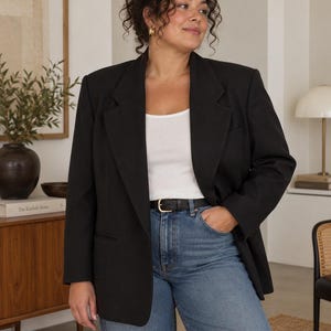 May include: A black blazer worn over a white tank top and blue denim jeans. The blazer features structured shoulders, notched lapels, and two front pockets. A thin black leather belt with a gold-toned buckle is worn around the waist of the jeans.