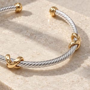 May include: A silver-tone twisted cable cuff bracelet featuring three polished gold-tone X-shaped accents spaced evenly along the band. The open-ended bracelet has small gold-tone spherical caps at each terminal end, creating a two-tone metal design.
